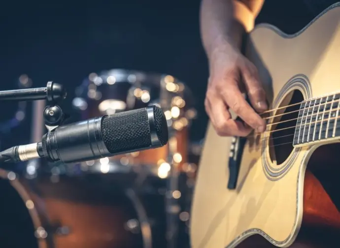 Male musician playing acoustic guitar behind microphone in recording studio.