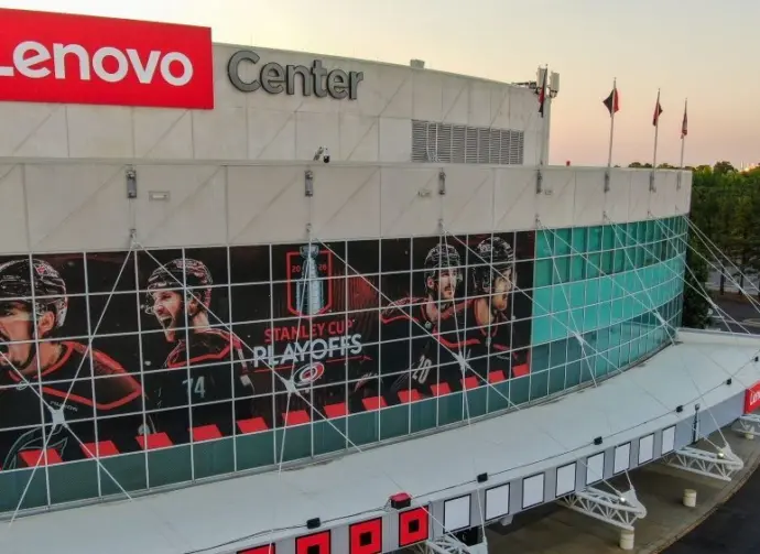 Aerial 4K drone shot captures a high-angle twilight view of the Lenovo Center arena^ home of the NHL's Carolina Hurricanes. The low golden hour sun rakes across the modern facade^ highlighting the reflective teal glass panels of the Carolina Hurricanes home venue before they face the Montreal Canadiens. The shot documents the wide^ geometric infrastructure layout of the empty parking lots^ main entrance gates^ and surrounding roadway logistics under a clean sunset gradient sky. Raleigh^ North Carolina^ USA - May 19^ 2026