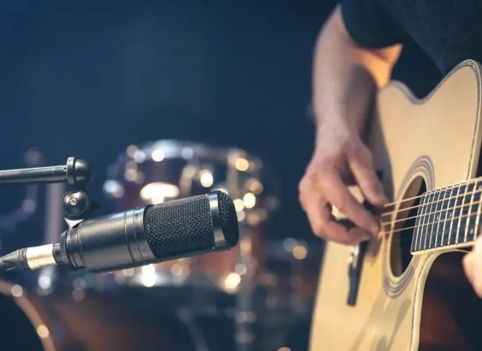 Male musician playing acoustic guitar behind microphone in recording studio.