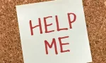 sticky note with 'help me' written in text in front of a cork board background