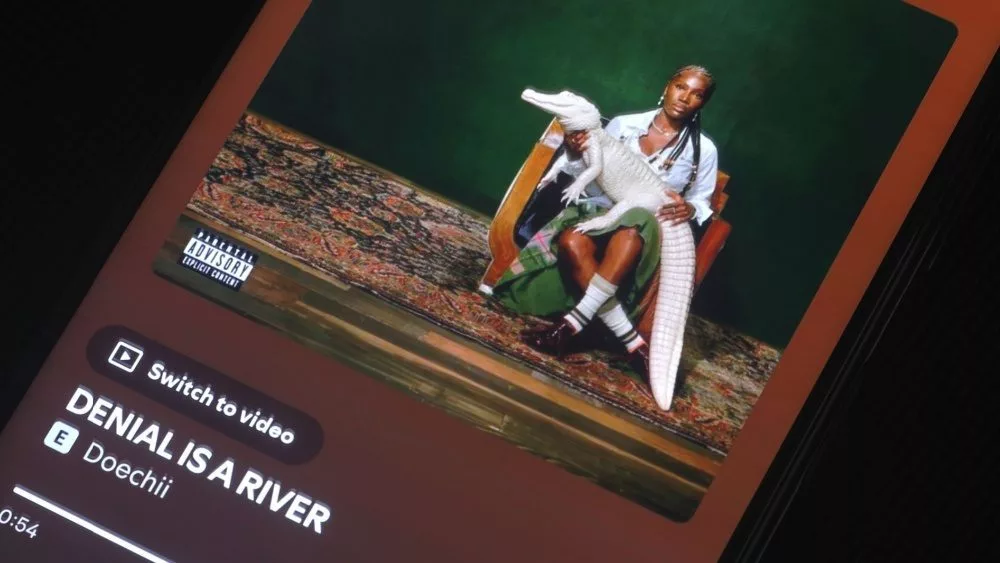 Album art of music artist Doechii from Spotify^ displayed on a phone screen on a dark surface^ playing song "DENIAL IS A RIVER"