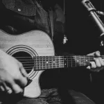 The guitarist plays an acoustic guitar with a capo in front of a microphone. The concept of music recording^ rehearsal or live performance.