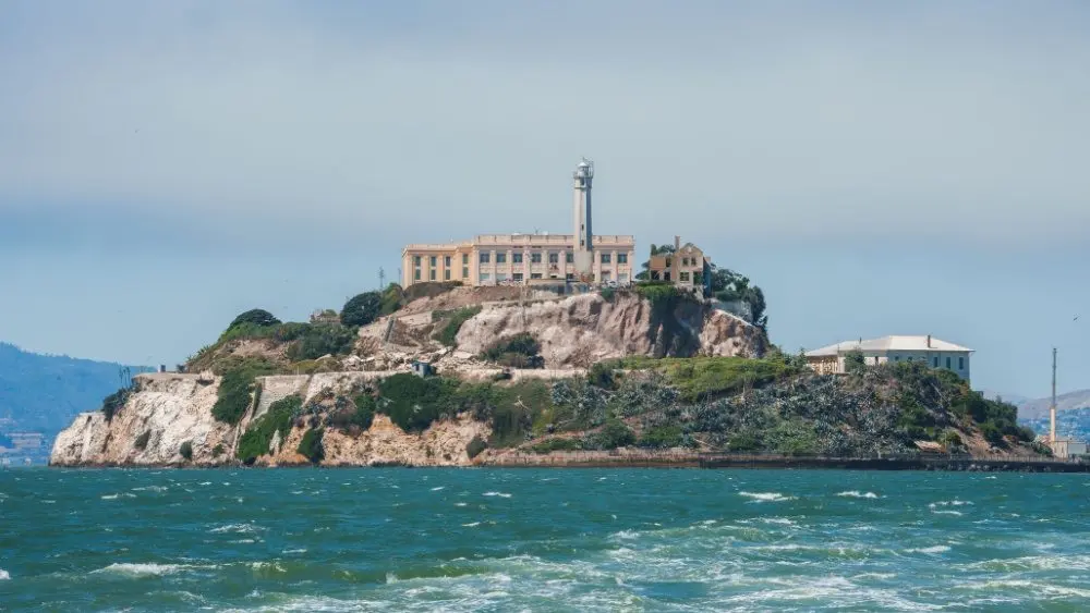 Alcatraz Island in San Francisco Bay^ California^ USA^ viewed from the water. Shows rugged terrain^ prison building^ lighthouse^ and choppy water under clear sky.