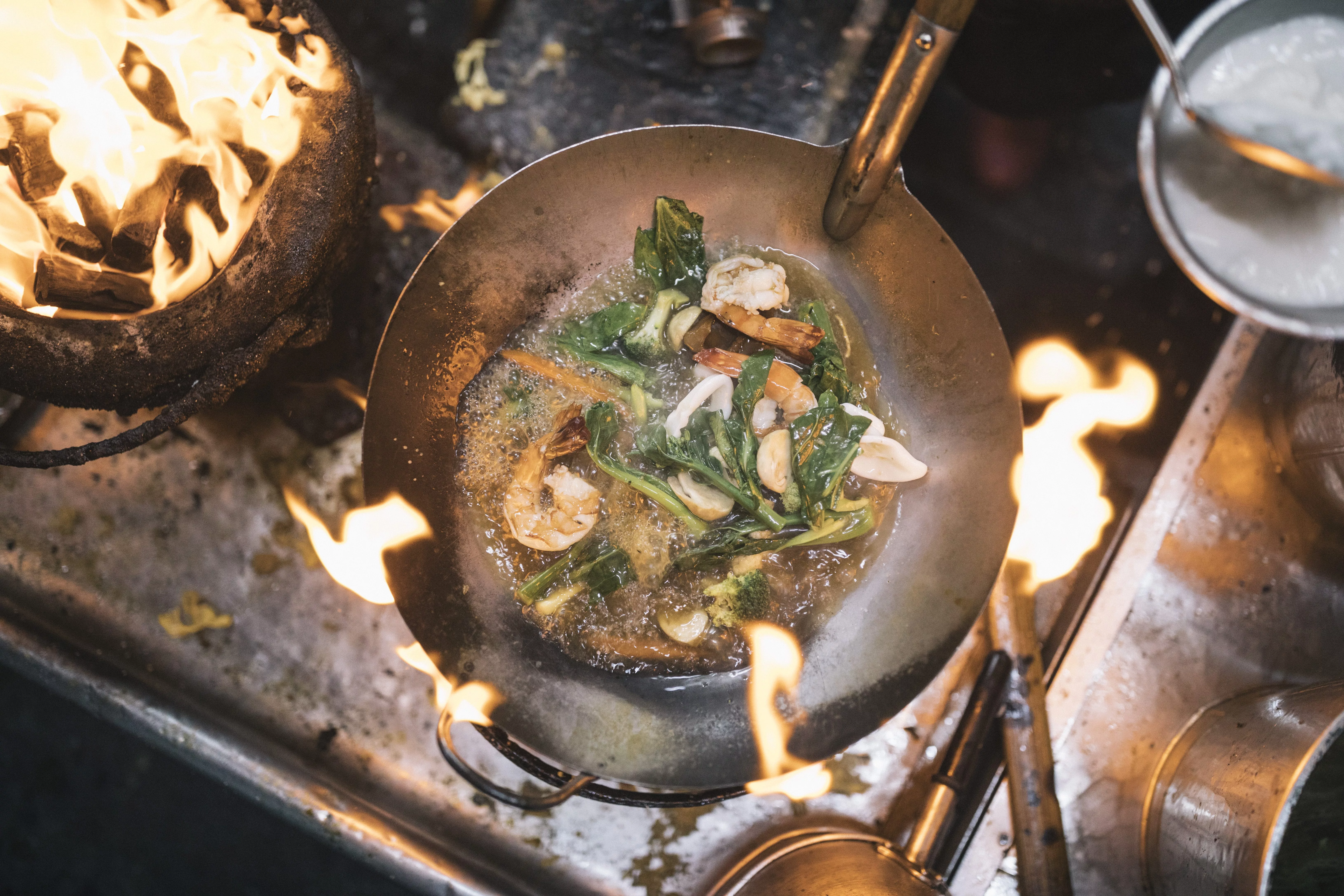 high-angle-close-up-of-stir-fry-with-prawns-and-green-vegetables-in-stainless-steel-wok-over-flame-of-charcoal-grill