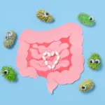 Paper Gut With Pills and Funny Bacteria on Blue Background: White pills arranged in a heart inside pink intestines with cartoon bacteria with googly eyes on blue background. Digestive health, probiotics
