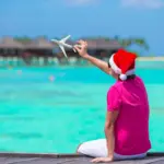 Young man in santa hat on white beach with miniature of airplane: Travel during the holidays