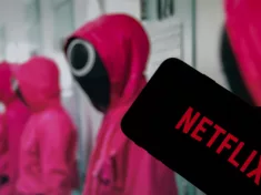 Netflix logo on the display of a smartphone in front of a television with the new series "Squid Game" ( focus on Netflix logo )