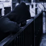 silhouette of man in dark hoodie outside leaning on fence