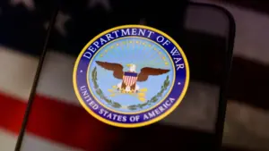 The United States Department of War seal is displayed on a smartphone screen. In the background^ an out-of-focus American flag is visible. This conceptual image illustrates themes of US military history^ national defense^ government institutions^ and historical conflicts.