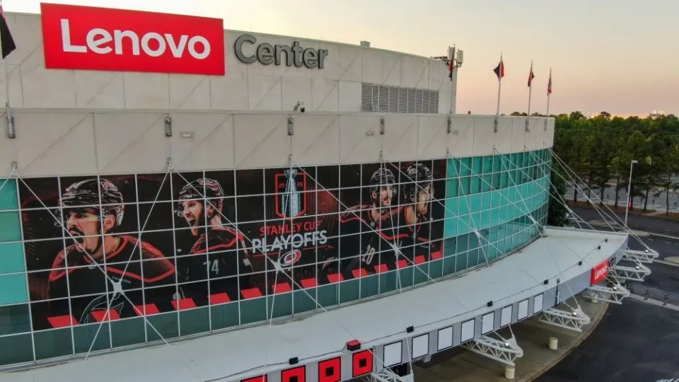 Aerial 4K drone shot captures a high-angle twilight view of the Lenovo Center arena^ home of the NHL's Carolina Hurricanes. The low golden hour sun rakes across the modern facade^ highlighting the reflective teal glass panels of the Carolina Hurricanes home venue before they face the Montreal Canadiens. The shot documents the wide^ geometric infrastructure layout of the empty parking lots^ main entrance gates^ and surrounding roadway logistics under a clean sunset gradient sky. Raleigh^ North Carolina^ USA - May 19^ 2026