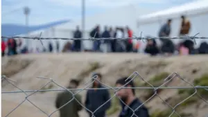 Barbed wire in refugee camp. Migrants behind chain link fence in camp. Group of people behind fence. Concept of prison^ freedom^ barrier^ security and migration. Refugees on their way to EU.