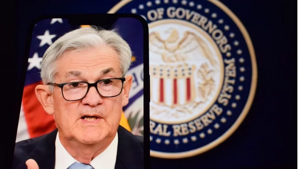 Federal Reserve (Fed) Chair Jerome Powell is visible on smartphone screen. In the background^ an inflation rate chart is displayed.