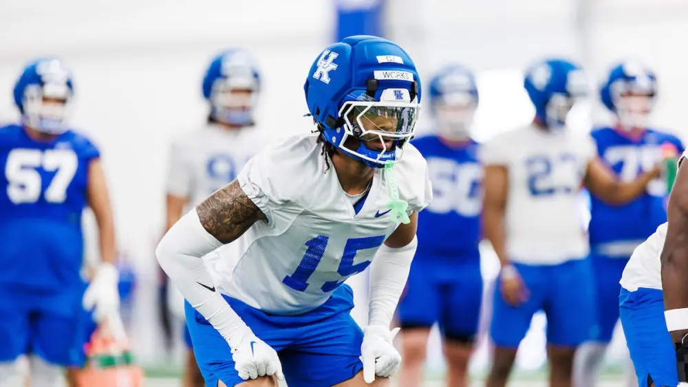 A football player wearing a blue helmet and jersey number 15 is in motion during a practice session, with teammates visible in the background.