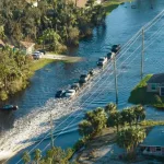 Hurricane flooded street with moving cars and surrounded with water houses in Florida residential area.