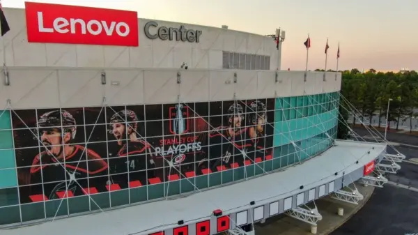 Aerial 4K drone shot captures a high-angle twilight view of the Lenovo Center arena^ home of the NHL's Carolina Hurricanes. The low golden hour sun rakes across the modern facade^ highlighting the reflective teal glass panels of the Carolina Hurricanes home venue before they face the Montreal Canadiens. The shot documents the wide^ geometric infrastructure layout of the empty parking lots^ main entrance gates^ and surrounding roadway logistics under a clean sunset gradient sky. Raleigh^ North Carolina^ USA - May 19^ 2026