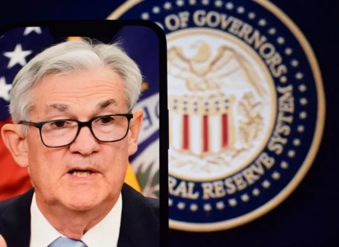 Federal Reserve (Fed) Chair Jerome Powell is visible on smartphone screen. In the background^ an inflation rate chart is displayed.