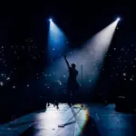 View from the stage stadium. Vocalist of a popular pop band on the background of the flashlights of phones during a concert. Fans switch on lights on smart phones on tribunes. Crowd waving cellphones