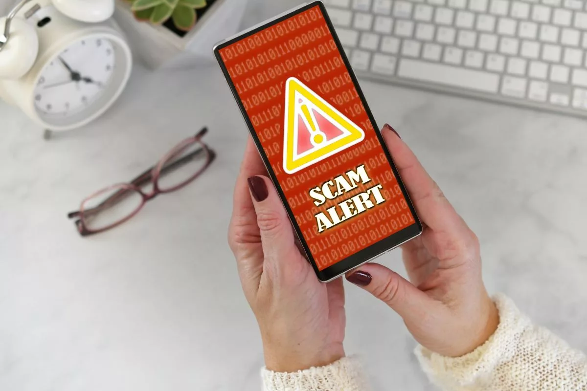 female-holding-a-phone-at-desk-with-scam-alert-warning-screen-in-red-and-yellow380329
