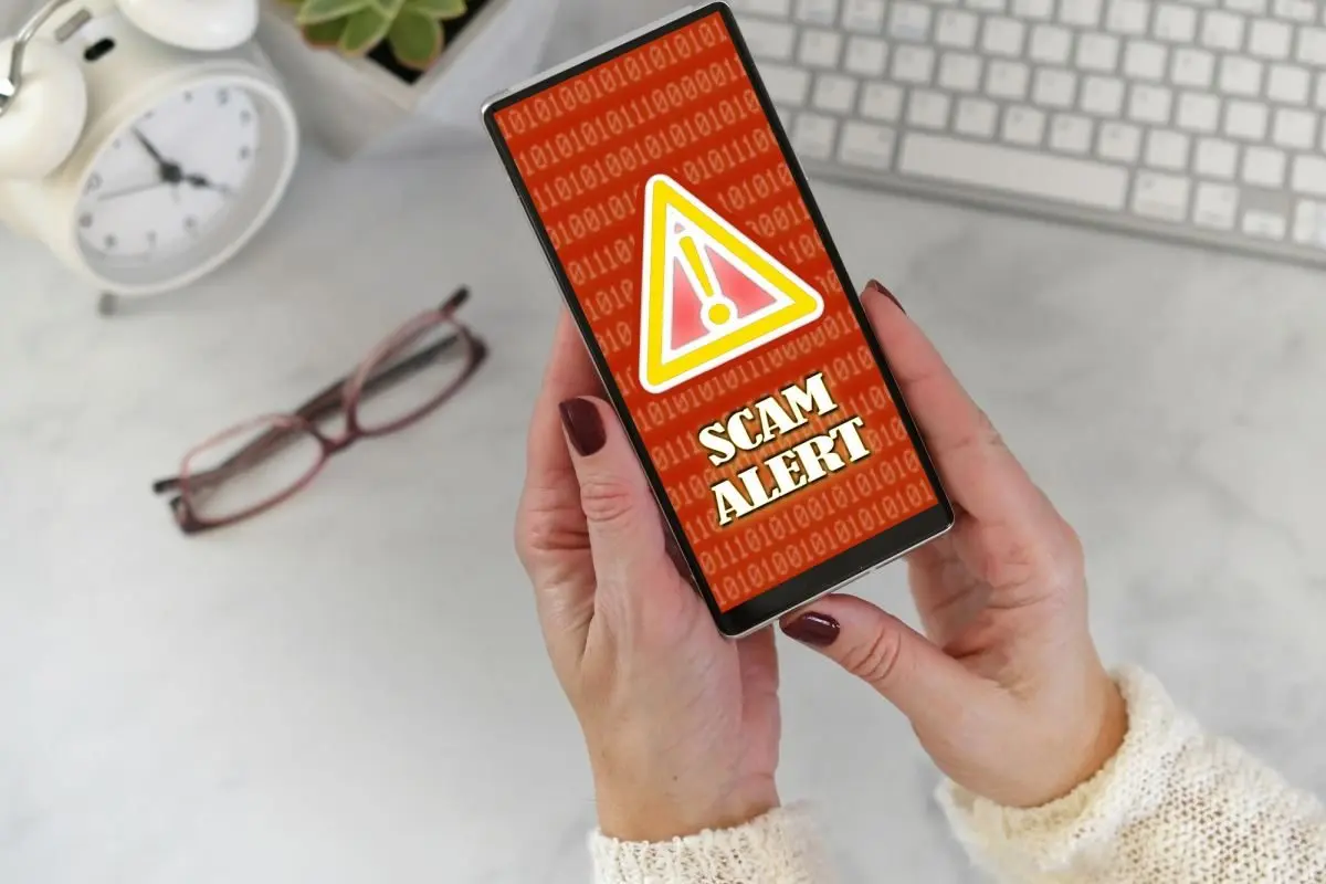 female-holding-a-phone-at-desk-with-scam-alert-warning-screen-in-red-and-yellow760366
