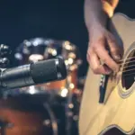 Male musician playing acoustic guitar behind microphone in recording studio.