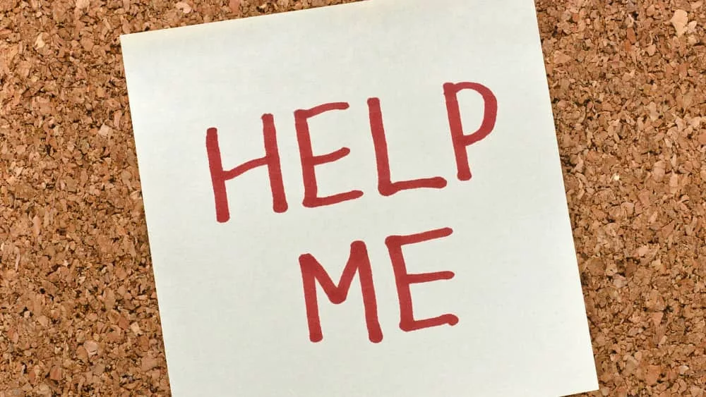 sticky note with 'help me' written in text in front of a cork board background