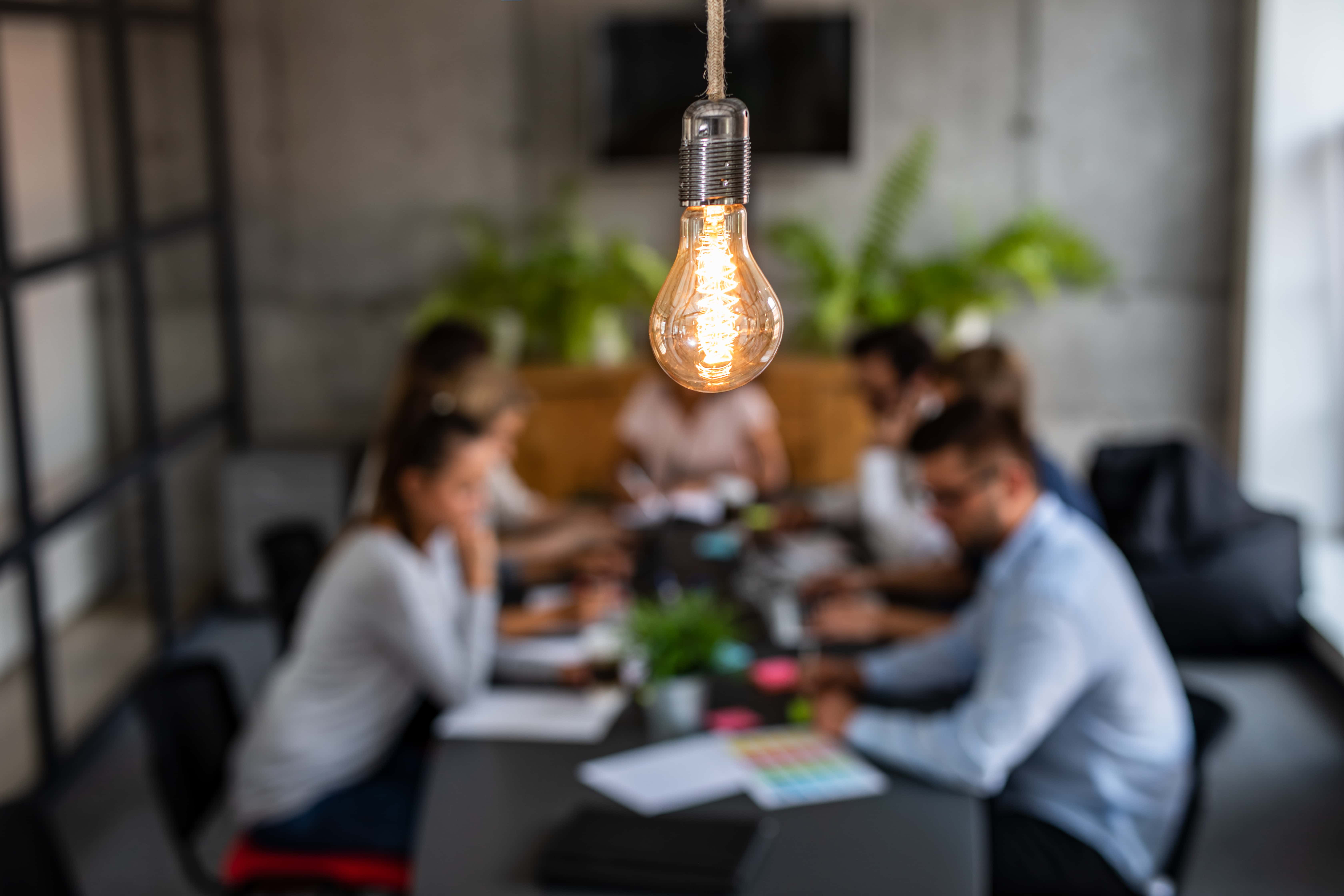 hanging lit lightbulb with blurred background of 5 people sitting around a conference room table with scattered papers on the table