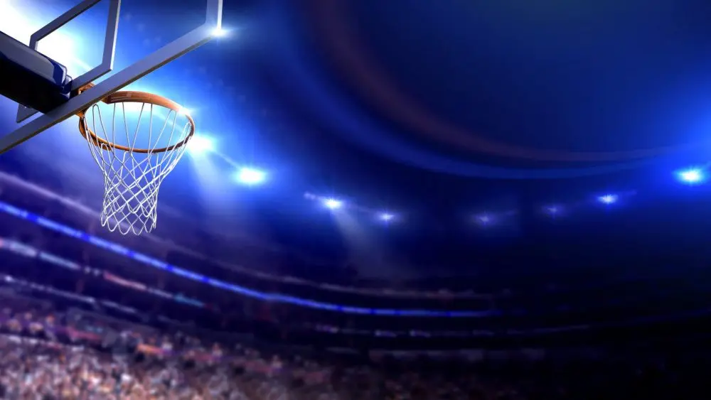 Basketball arena view of basketball hoop in dimmed arena^ crowd in silhouette^ lights turned on brightly