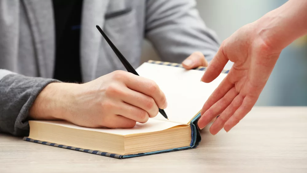 author-signing-autograph-in-own-book-at-wooden-table-on-light-bl