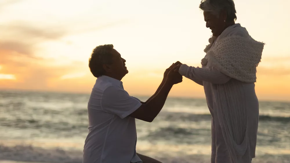 side-view-of-senior-biracial-man-holding-hands-with-woman-while-kneeling-and-proposing-at-beach-lifestyle-love-and-copy-space