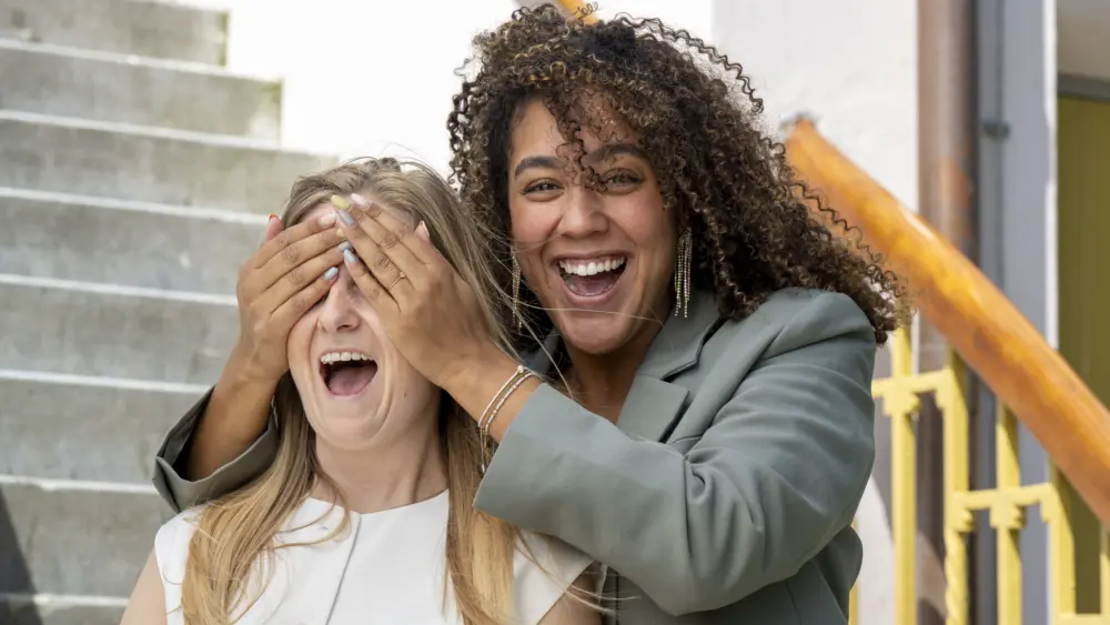woman-covers-friends-eyes-with-her-hands-as-they-both-laugh-on-outdoor-steps