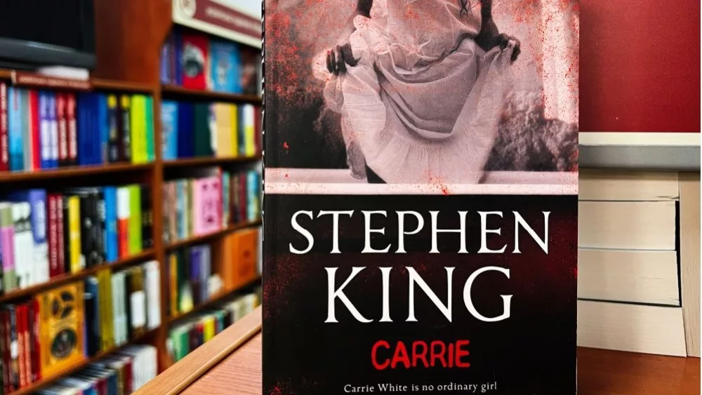 Paperback cover of Carrie by Stephen King shown on a bookshop shelf background