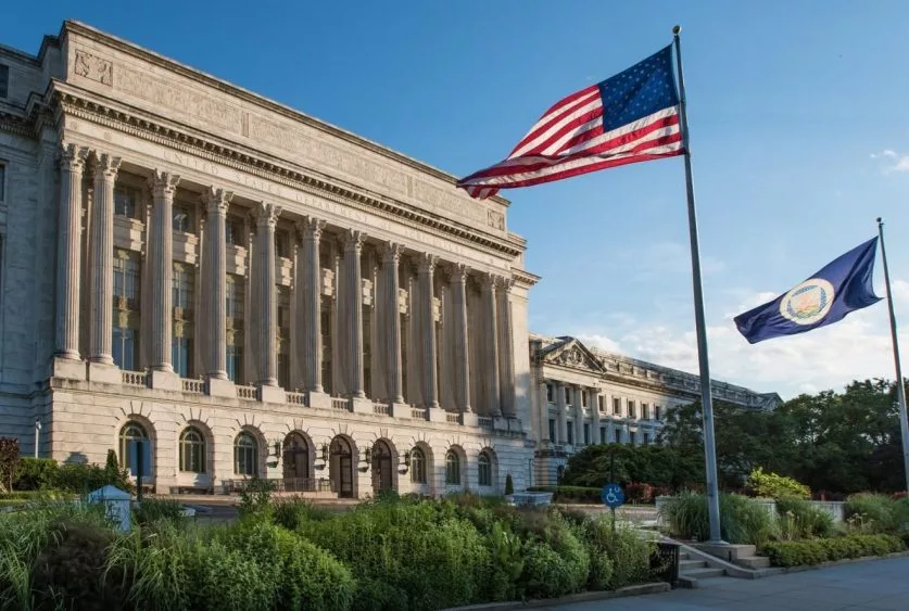 The image shows the United States Department of Agriculture building with columns^ featuring the American flag and USDA flag in front^ set against a clear blue sky.