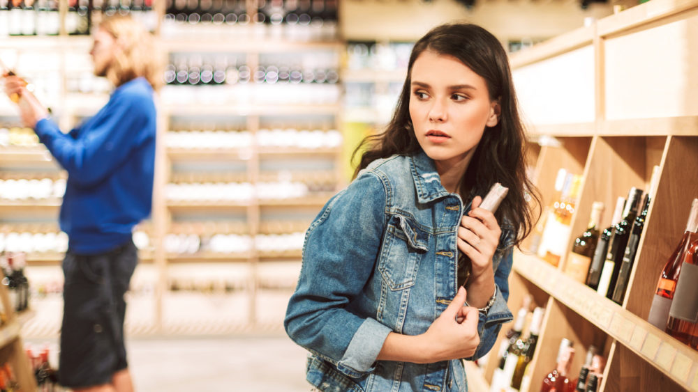young-frightened-woman-in-denim-jacket-trying-steal-bottle-of-wine-in-modern-supermarket