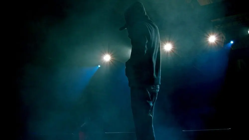 Rap artist performing on stage under soffit lights. Concert backlighting and illumination during live music show. Singer in hoodie holding microphone^ starting the concert