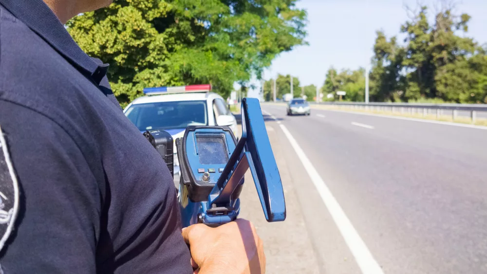 policeman-holding-laser-speed-gun-near-police-car-on-highway-background-selective-focus-part-of-body