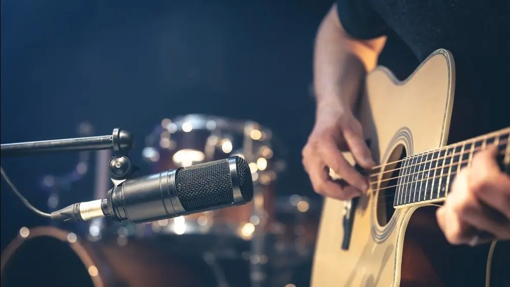 Male musician playing acoustic guitar behind microphone in recording studio.