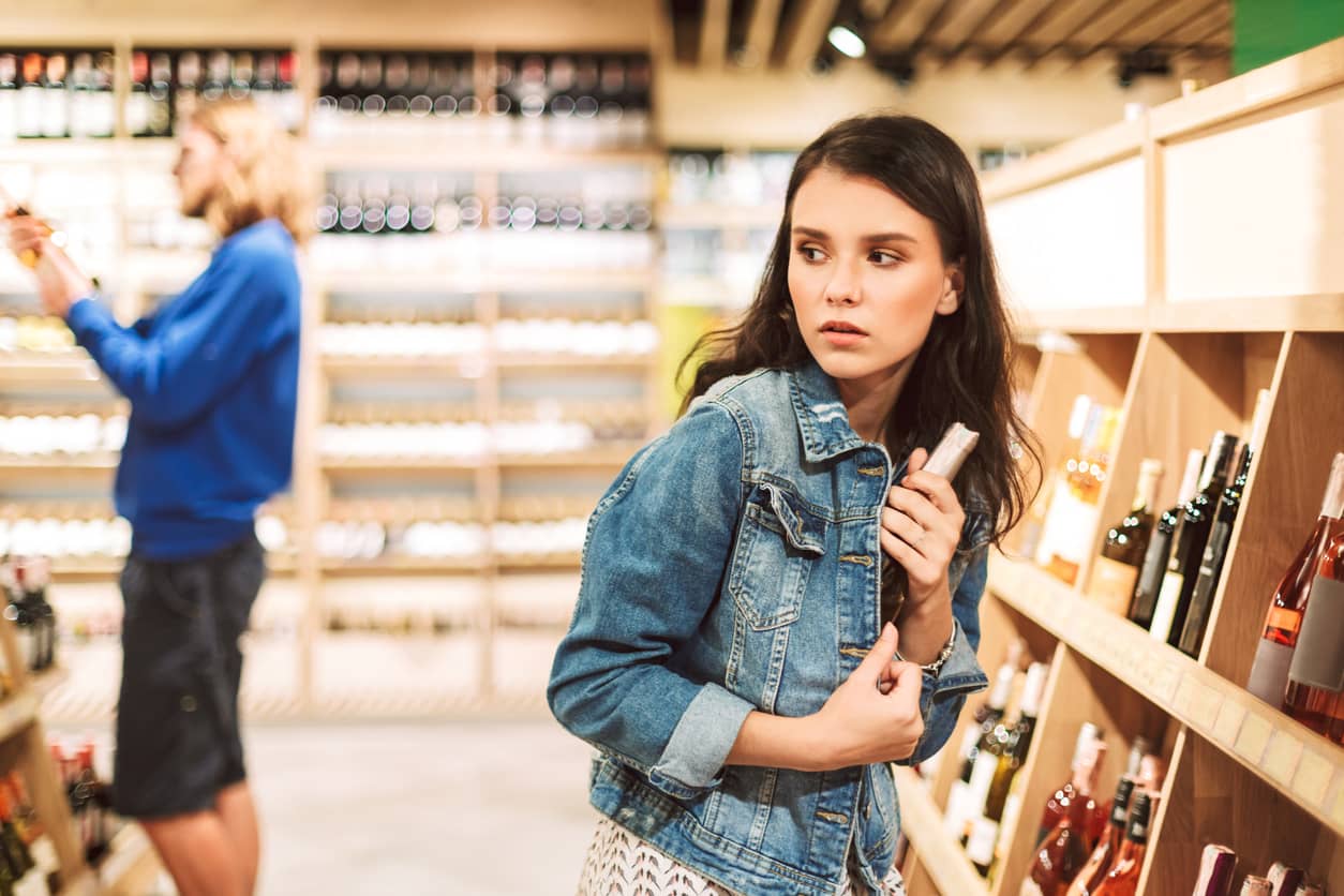 young-frightened-woman-in-denim-jacket-trying-steal-bottle-of-wine-in-modern-supermarket