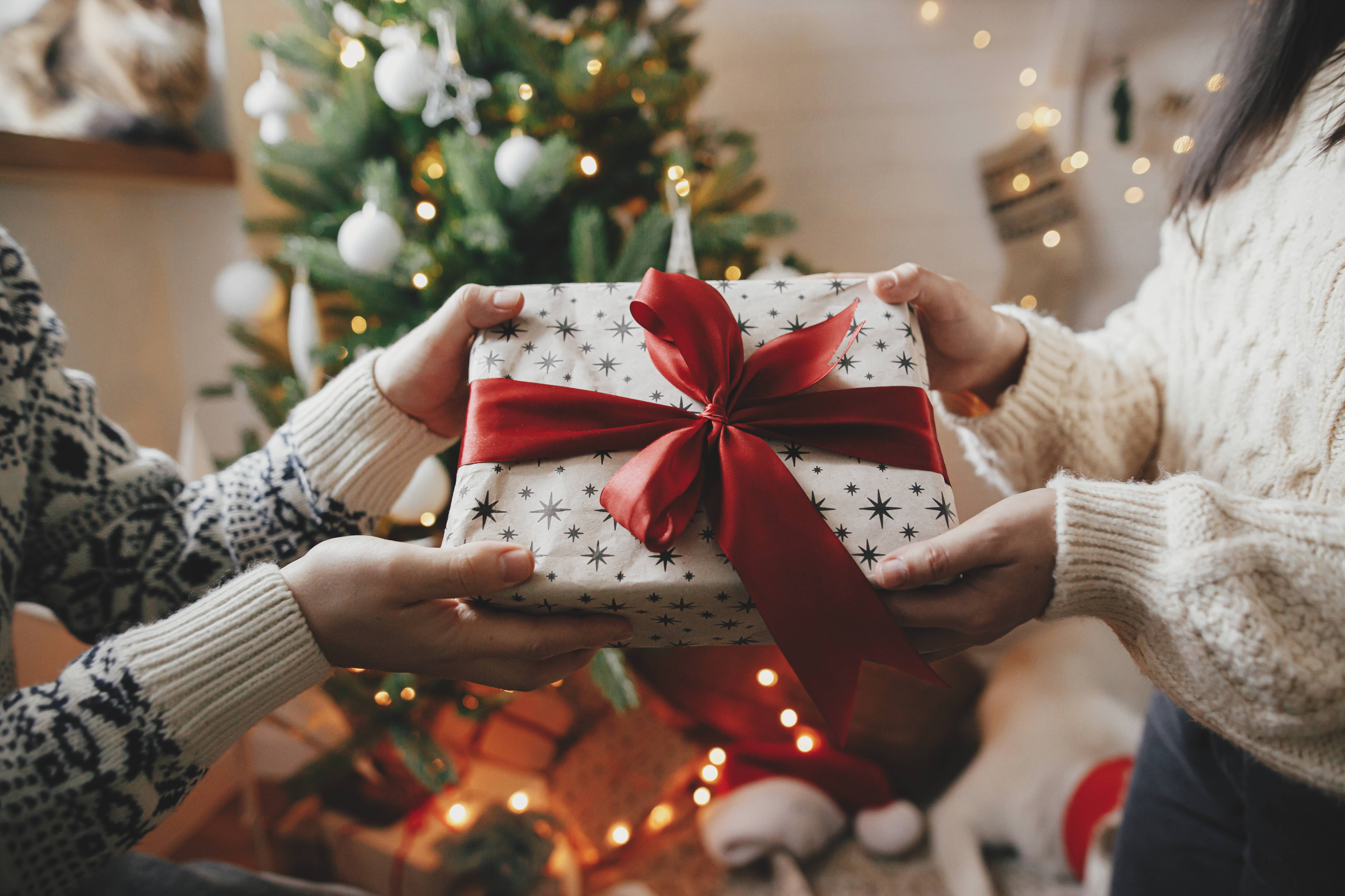 couple-exchanging-christmas-gift-with-red-bow-on-background-of-christmas-tree-with-lights-stylish-couple-hands-holding-present-with-red-ribbon-close-up-in-festive-decorated-room-happy-holidays