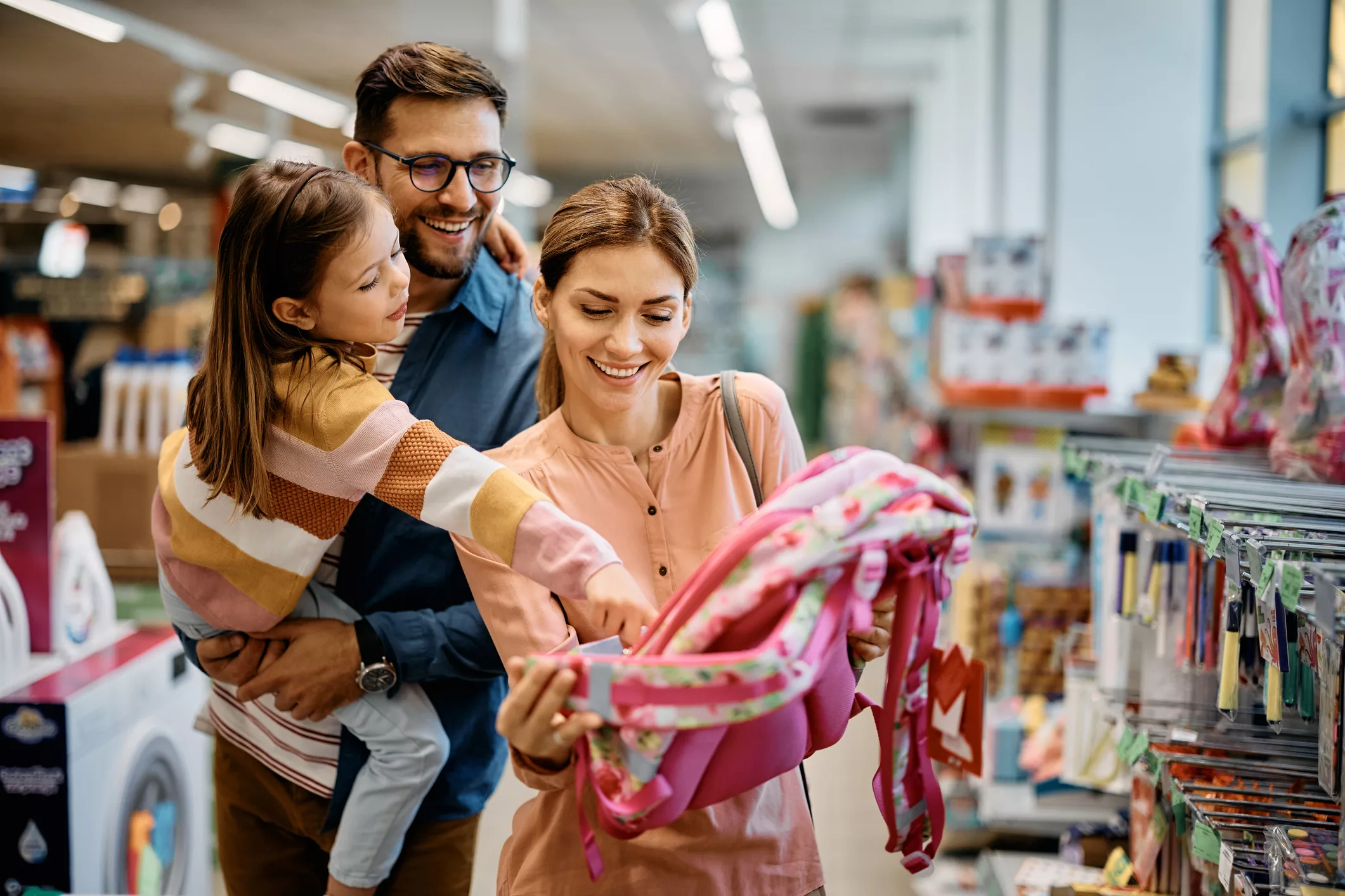 happy-little-girl-pointing-at-backpack-while-buying-school-supplies-with-her-parents-in-supermarket
