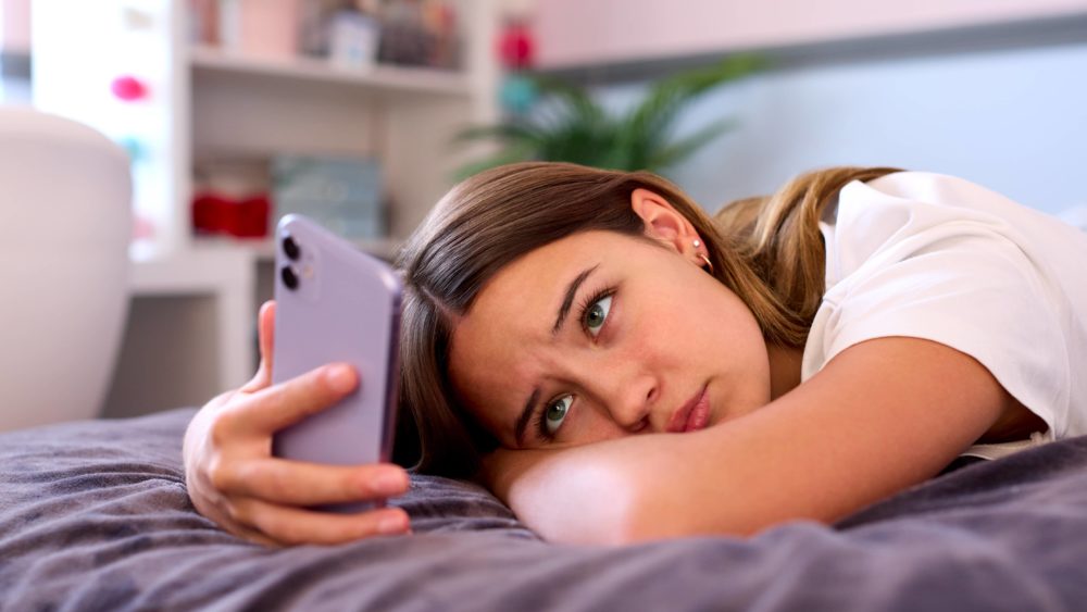 depressed-teenage-girl-lying-on-bed-at-home-looking-at-mobile-phone