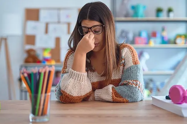 young-beautiful-teacher-woman-wearing-sweater-and-glasses-sitting-on-desk-at-kindergarten-tired-rubbing-nose-and-eyes-feeling-fatigue-and-headache-stress-and-frustration-concept