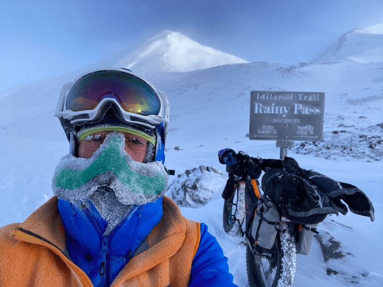 1. Peter Delamere poses in front of a sign in Rainy Pass in the Alaska Range during the 2024 Iditarod Trail Invitational race. On his face is a “nose-hat” invented by Fairbanks athlete Shalane Frost. Photo by Peter Delamere.
