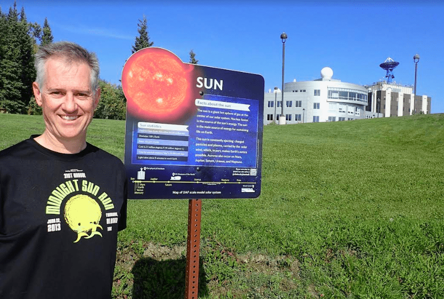 Peter Delamere pauses in front of a UAF Planet Walk sign at the University of Alaska Fairbanks, with the International Arctic Research Center and the Geophysical Institute in the background. Photo by Ned Rozell.
