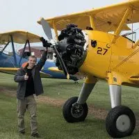 Brett Anderson giving a thumbs up beside a yellow Stearman biplane with its propeller visible