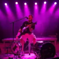 Sean Kistler sings and plays acoustic guitar on a stage lit by purple spotlights^ with a kick drum bearing his name in front of him