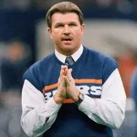 Mike Ditka in a navy Bears sweater vest and tie^ palms pressed together^ on the sideline