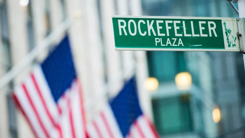 Close-up view of green street sign depicting it is Rockefeller Plaza in Midtown Manhattan^ New-York. Blurred American flags in the background