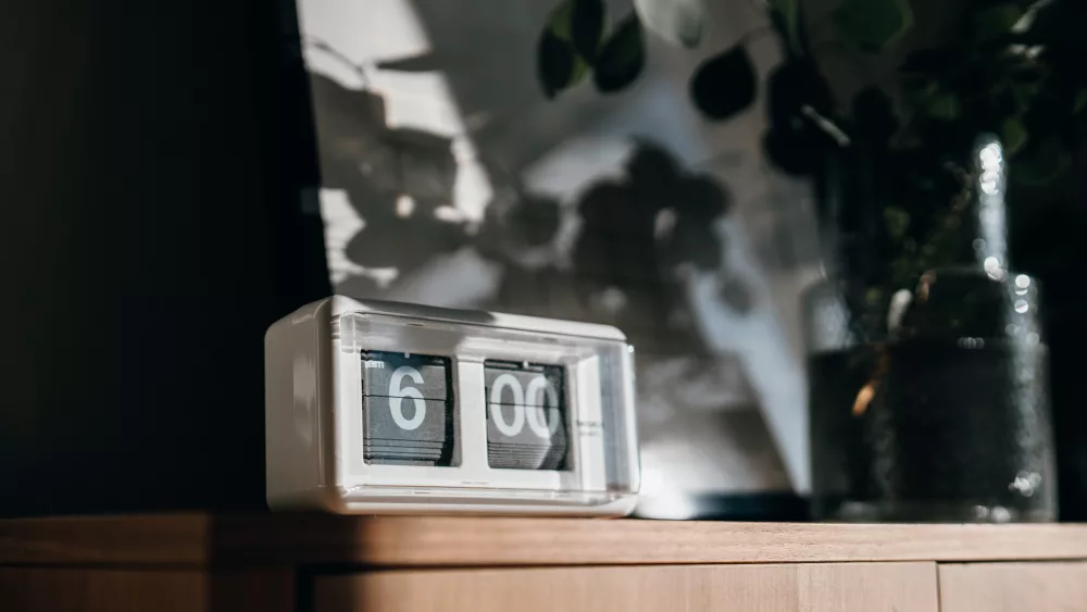 a-white-digital-flip-clock-shows-six-oclock-on-a-brown-wooden-cabinet-next-to-a-fresh-eucalyptus-plant-in-the-living-room-with-sunbeam-shining-through-the-window-on-a-fresh-beautiful-morning-a-bran