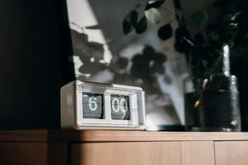 a-white-digital-flip-clock-shows-six-oclock-on-a-brown-wooden-cabinet-next-to-a-fresh-eucalyptus-plant-in-the-living-room-with-sunbeam-shining-through-the-window-on-a-fresh-beautiful-morning-a-bran