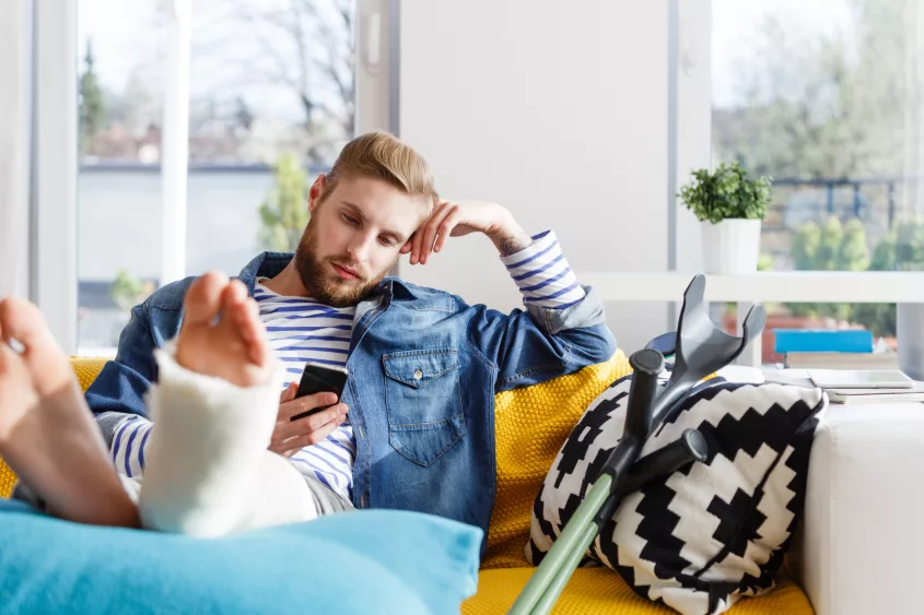 young-man-with-broken-leg-using-smart-phone-at-home